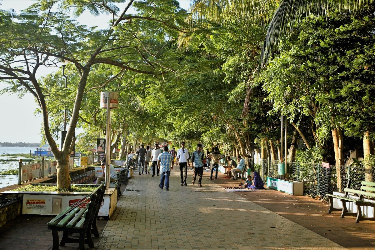 Foto Marine Drive di Kochi; orang-orang berjalan di bawah naungan pepohonan.